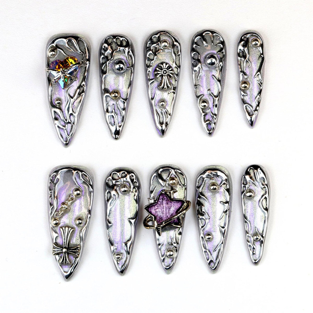 A set of ten silver-toned, stiletto-shaped press-on nails. Each nail features intricate, raised designs and embellishments, including crystals, chains, and star-shaped accents. The nails have a pearlescent, iridescent finish.