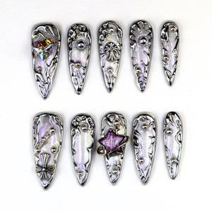 A set of ten silver-toned, stiletto-shaped press-on nails. Each nail features intricate, raised designs and embellishments, including crystals, chains, and star-shaped accents. The nails have a pearlescent, iridescent finish.