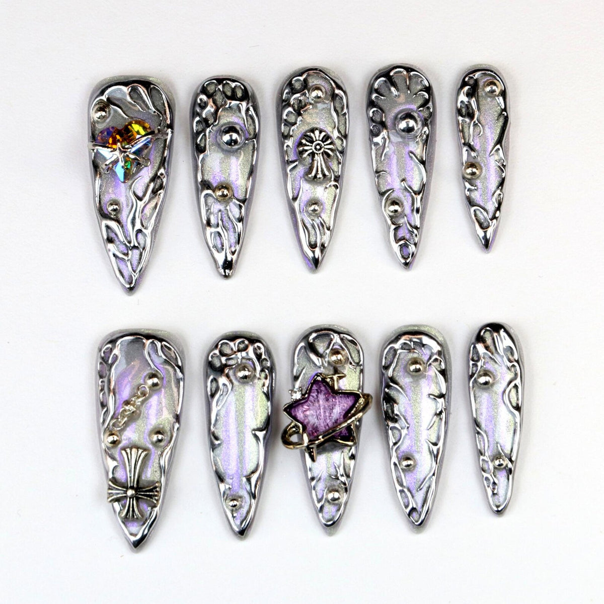A set of ten long, silver-toned artificial nails. Each nail features a pearlescent, iridescent finish with ornate, raised silver designs and decorative embellishments, including gems and crosses. The nails are arranged on a white surface.