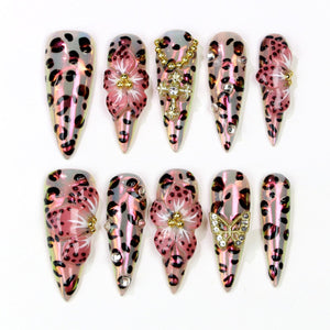 A set of ten long, pointed artificial nails. Each nail features a pink and black leopard print design. Some nails are adorned with pink floral embellishments, rhinestones, and gold-colored charms, including a butterfly and a cross.