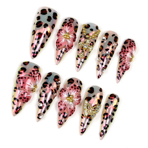 A collection of ten long, pointed artificial nails. The nails feature a variety of designs, including leopard print, pink floral accents, and gold or crystal embellishments. The nails have a glossy, iridescent finish, and are arranged on a white back