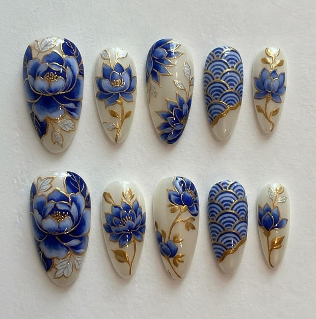 Set of ten oval-shaped false nails. Each nail is painted with a cream base and features intricate designs in blue and gold. Designs include floral patterns and wave-like motifs, with gold accents.