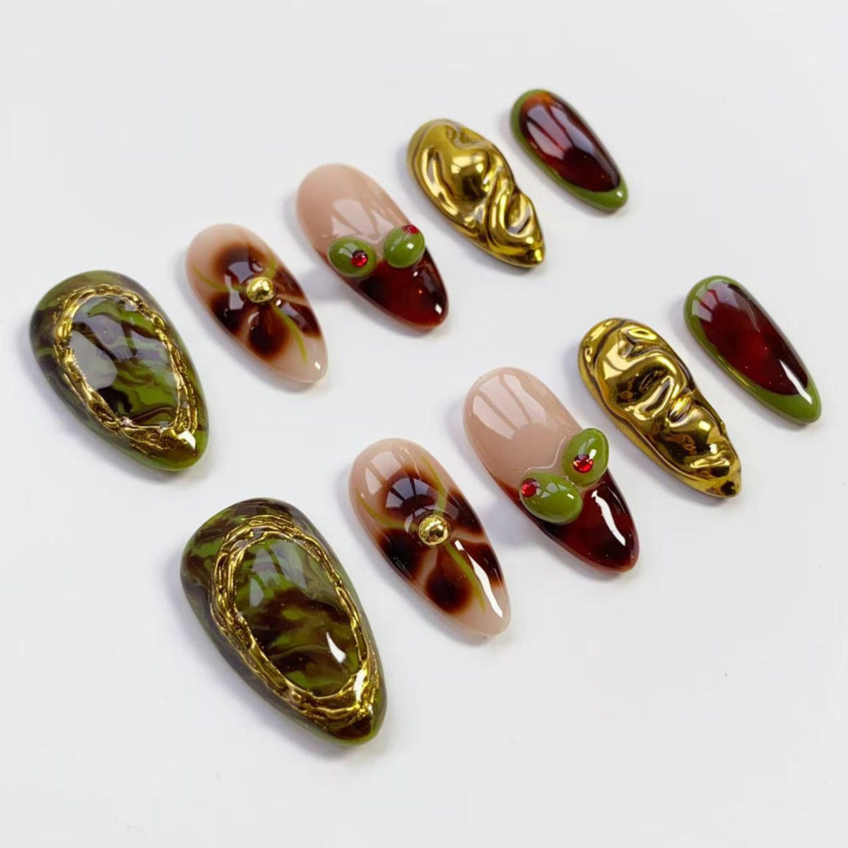 A collection of artificial nails in various designs. Some nails feature a gold metallic border with green and brown marbling. Others have a nude base with brown floral patterns or red tips with green olive-shaped accents. Some nails are gold, texture