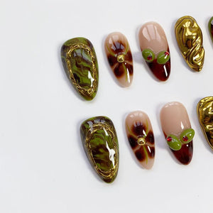 A collection of artificial nails in various designs. Some nails feature a green and brown marbled effect with gold trim. Others have a nude base with brown and gold accents, and some have olive-like embellishments.