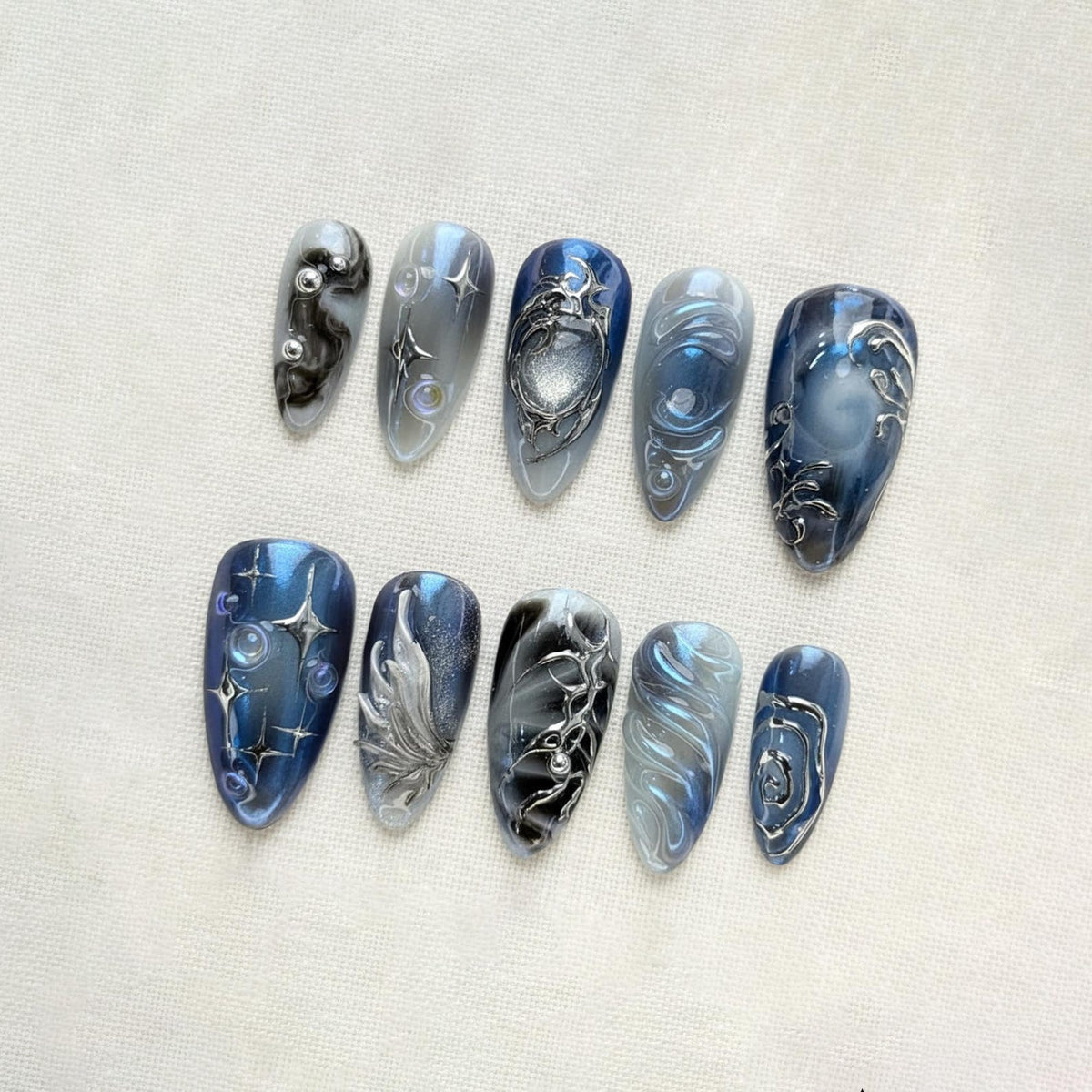 A set of ten almond-shaped false nails. Each nail features a unique design with shades of blue, gray, and silver. Some nails have swirling patterns, while others showcase intricate silver embellishments and star-like accents.