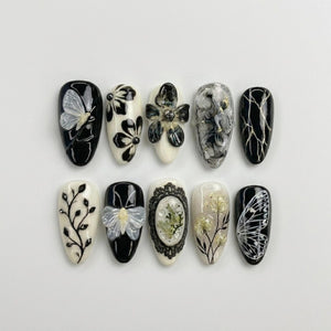 A set of ten press-on nails featuring a black and white color palette. Designs include a translucent butterfly, floral patterns, marbled textures, intricate lace-like frames, botanical sprigs, and delicate wing motifs with gold accents.