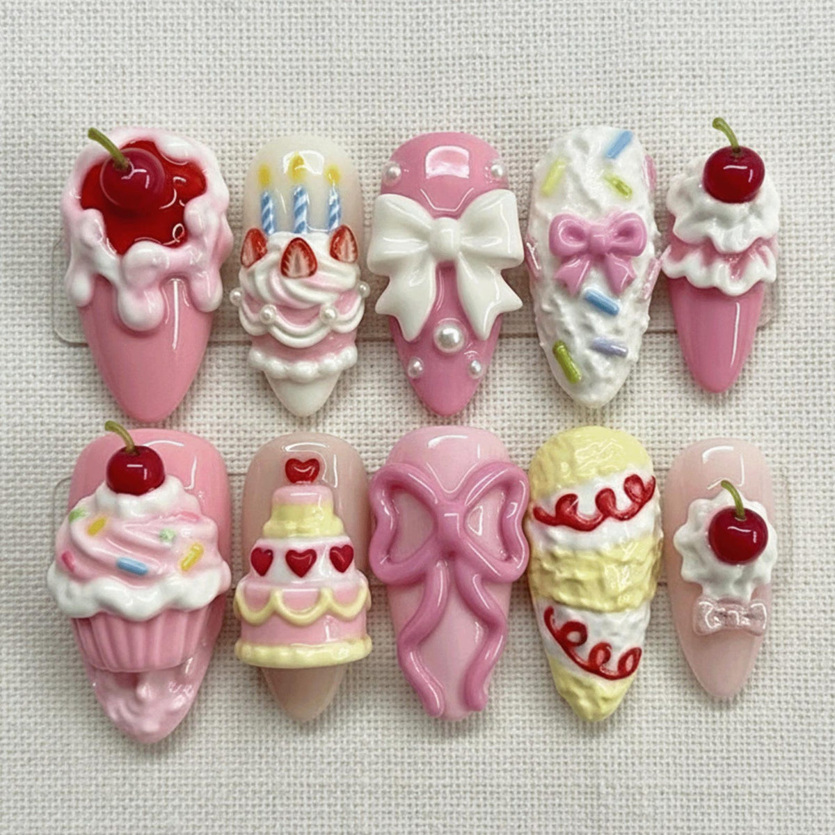A collection of ten artificial nails with various dessert-themed designs. The nails feature pink, white, and yellow colors, with elements like cherries, bows, and cake decorations. Each nail has a unique design, including cupcakes and birthday cakes.