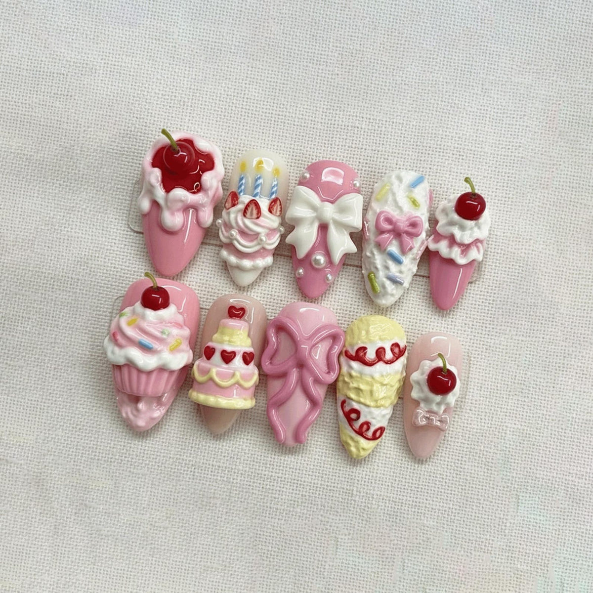 A collection of ten artificial nails, each uniquely designed with a dessert theme. The nails feature pink, white, and yellow colors, with designs including cherries, bows, cupcakes, and ice cream cones. The nails are arranged on a white surface.