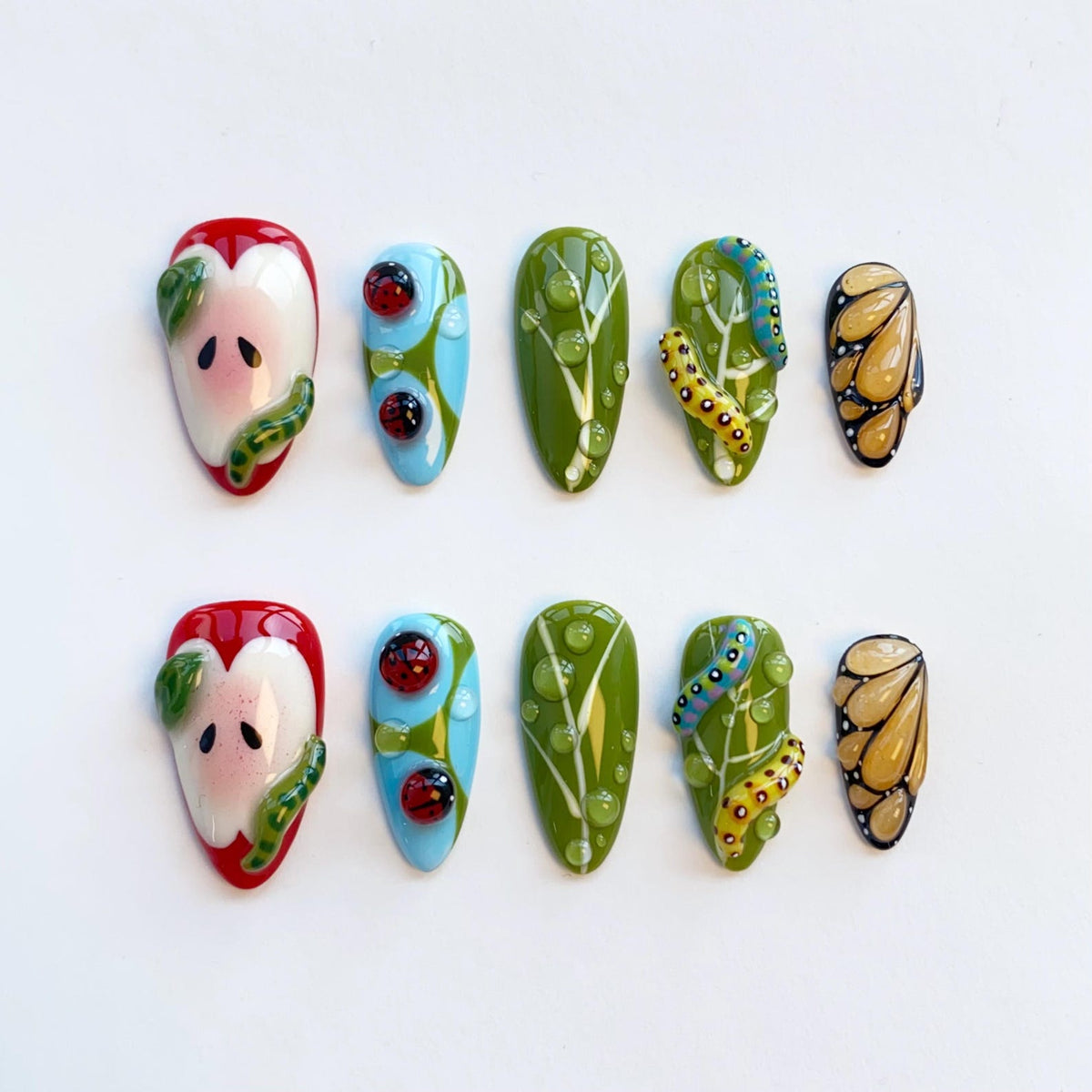 A set of ten artificial nails with unique designs. The designs include a red apple, ladybugs, green leaves with water droplets, a caterpillar, and a butterfly wing. The nails are painted in various colors, including red, green, blue, yellow, and whit