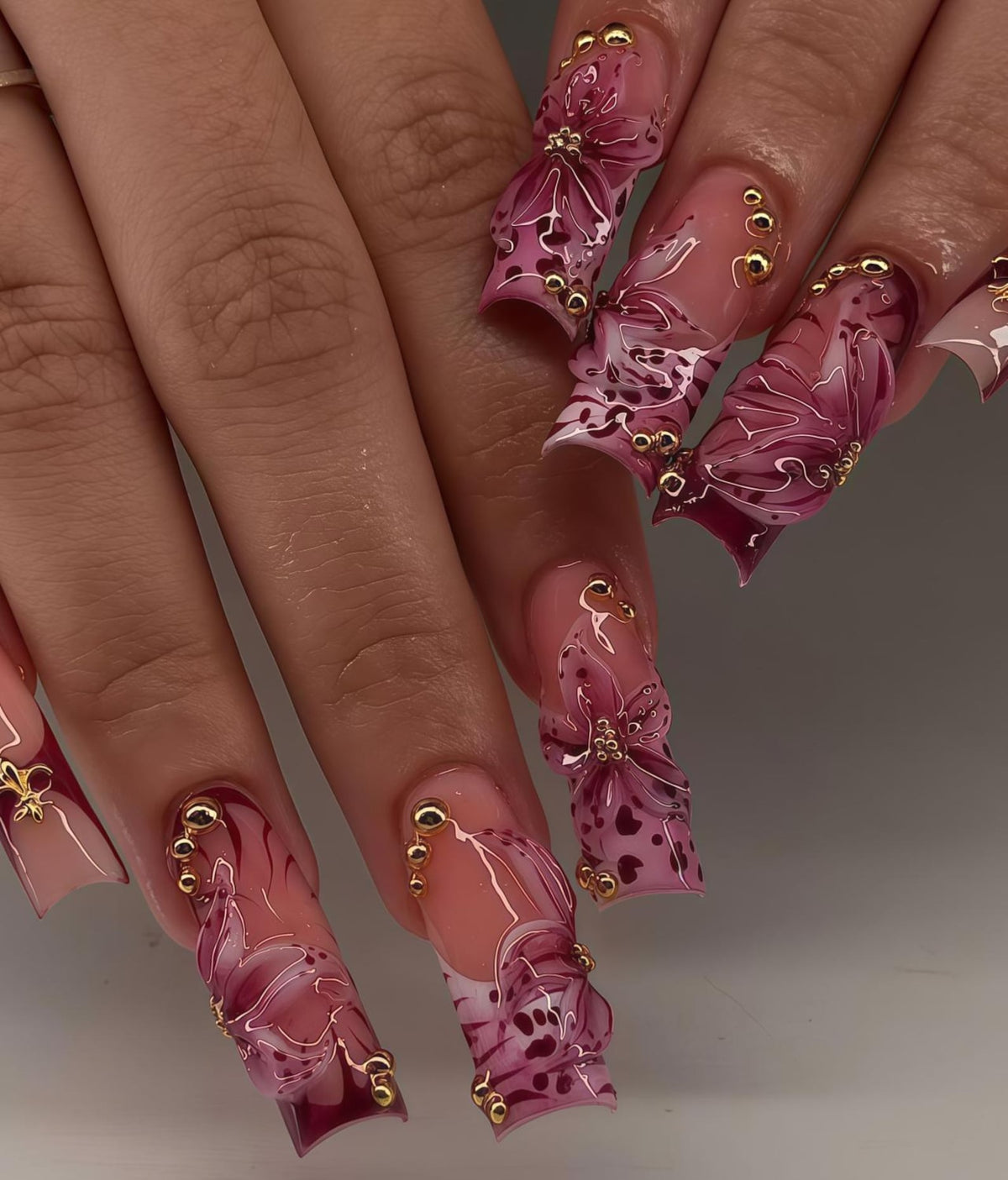 Long, sculpted acrylic nails with a gradient pink base, adorned with 3D floral designs in shades of pink and burgundy. Gold embellishments add a touch of glamour to the nail art.