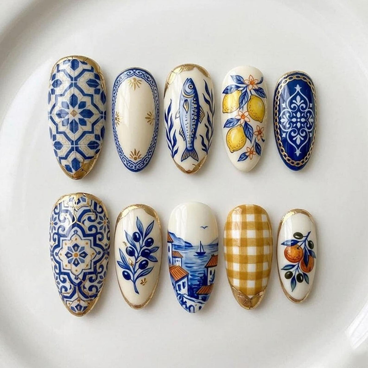 A set of ten almond-shaped nail art designs. Each nail features a unique hand-painted design, including blue and white tile patterns, a fish, lemons, olives, a coastal scene, and a gingham pattern. Gold trim accents each nail.