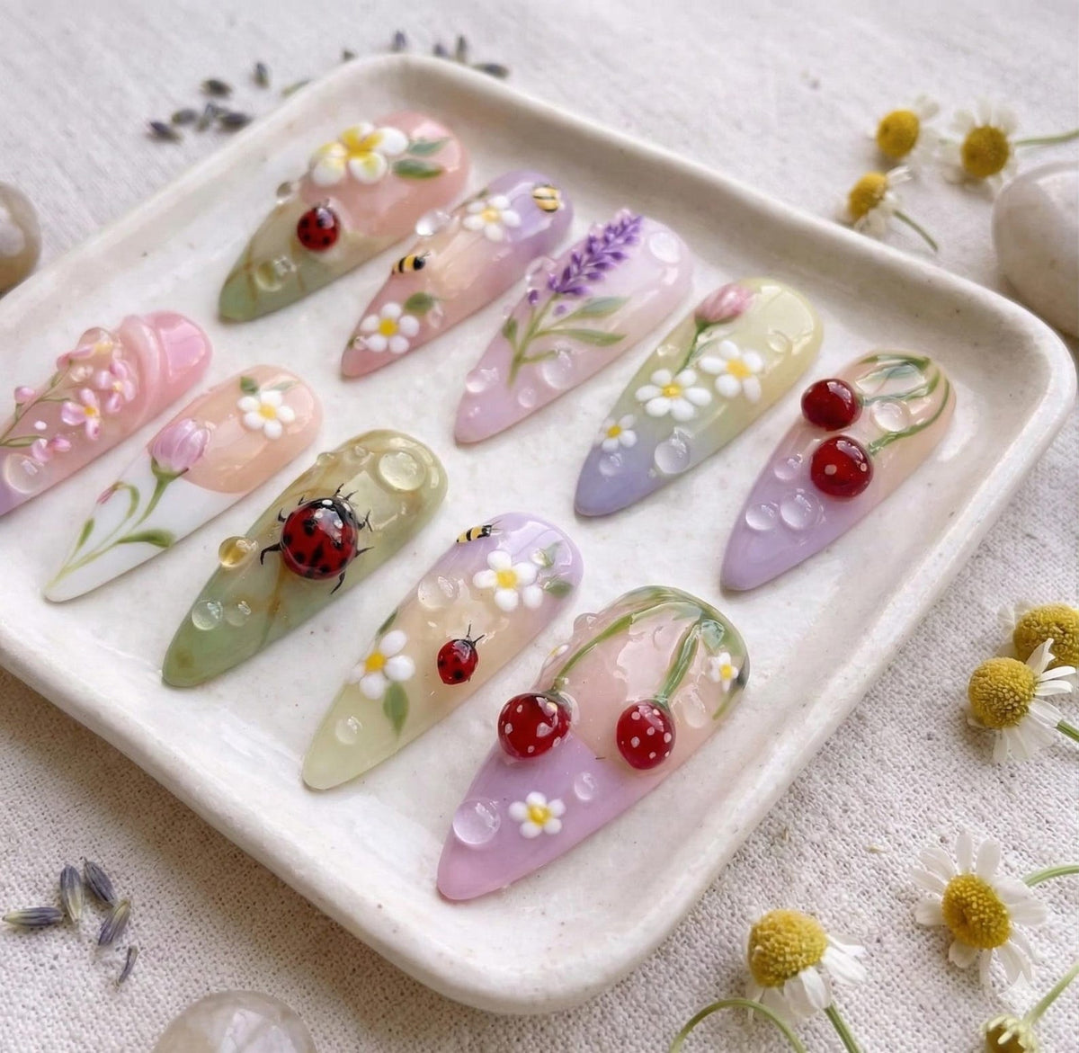 A collection of ten almond-shaped acrylic nails, each uniquely designed with floral, insect, and fruit motifs. The nails feature a variety of colors, including pink, green, and purple, with detailed artwork of ladybugs, daisies, lavender, and cherrie