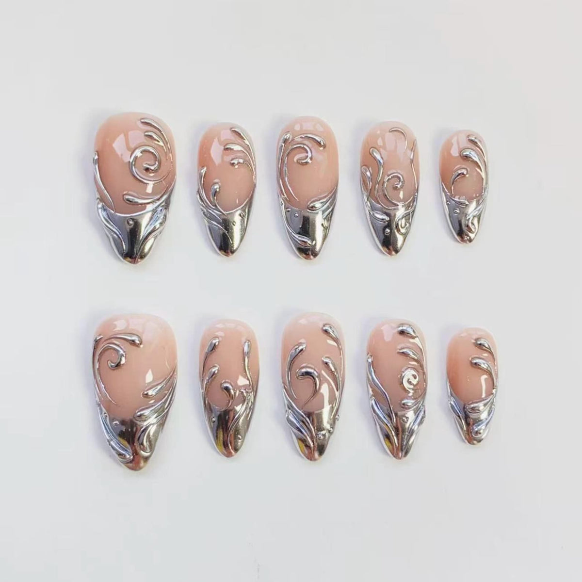 Set of ten artificial nails in various sizes. Each nail features a nude-colored base with a silver metallic design. The silver design includes a swirl pattern and a metallic tip, creating a decorative and elegant look.