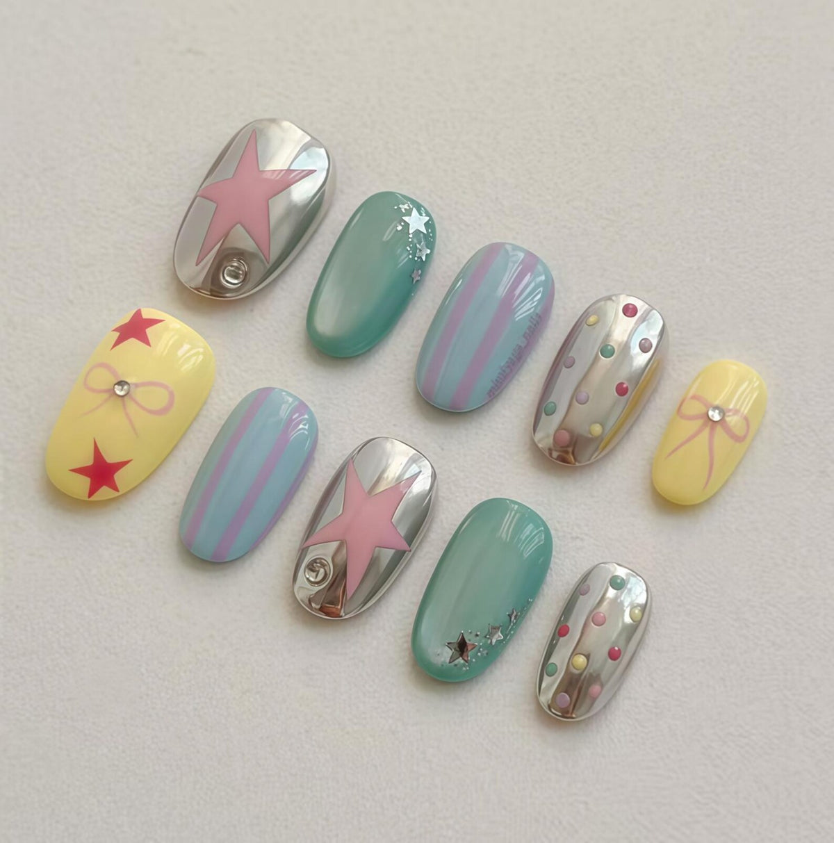 A set of ten artificial nails in various designs. The nails feature a mix of colors including silver, yellow, and teal. Some nails have star and bow designs, while others have stripes and polka dots. Each nail is oval-shaped.