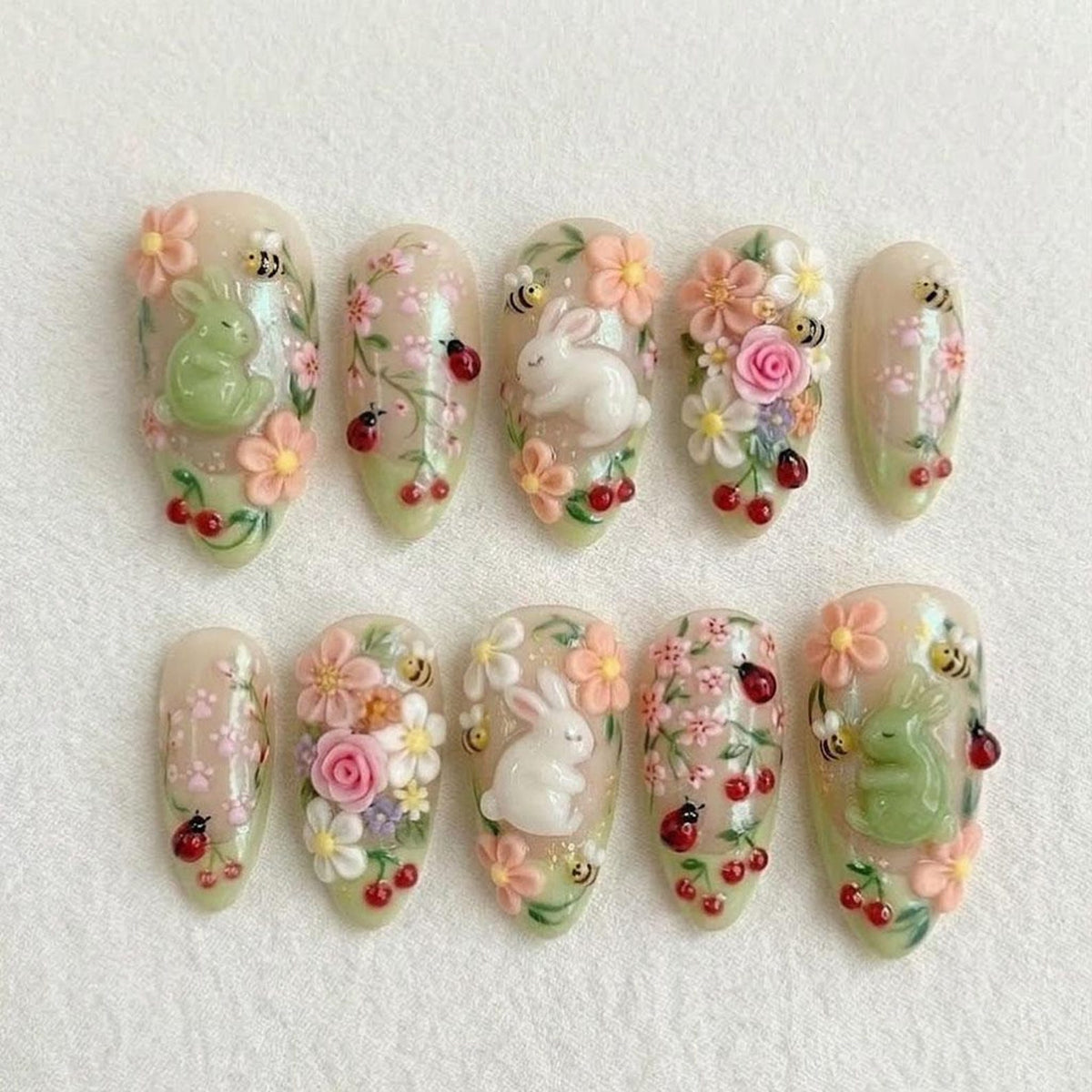 A set of ten almond-shaped artificial nails. Each nail features a spring-themed design with 3D elements, including white rabbits, pink and white flowers, red cherries, ladybugs, and bees. The nails have a light green base.