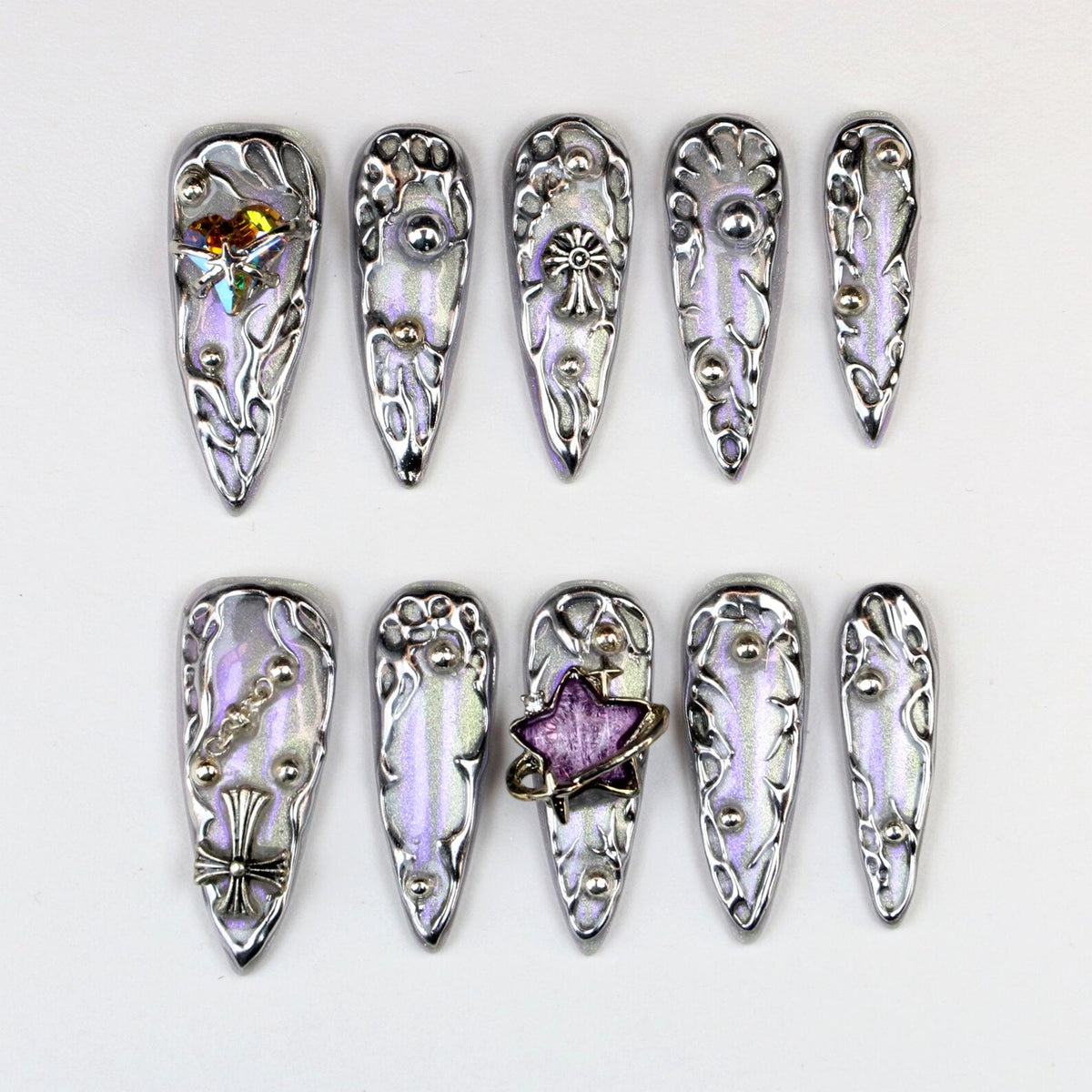 Set of ten silver and iridescent false nails. Each nail features a unique design with decorative elements like studs, crosses, and a star. The nails have a pointed shape and are arranged on a white surface.