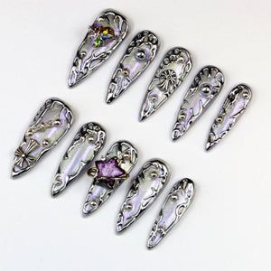 A collection of silver-toned, ornate nail art designs. Each nail features intricate detailing, including swirling patterns, small embellishments, and some with colorful accents. The nails are arranged on a white surface.