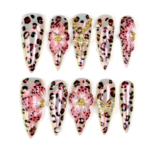 A set of ten long, pointed artificial nails. The nails feature a leopard print design with pink and brown spots. Some nails are adorned with 3D pink flowers, rhinestones, and gold-colored charms, including a butterfly and a cross.