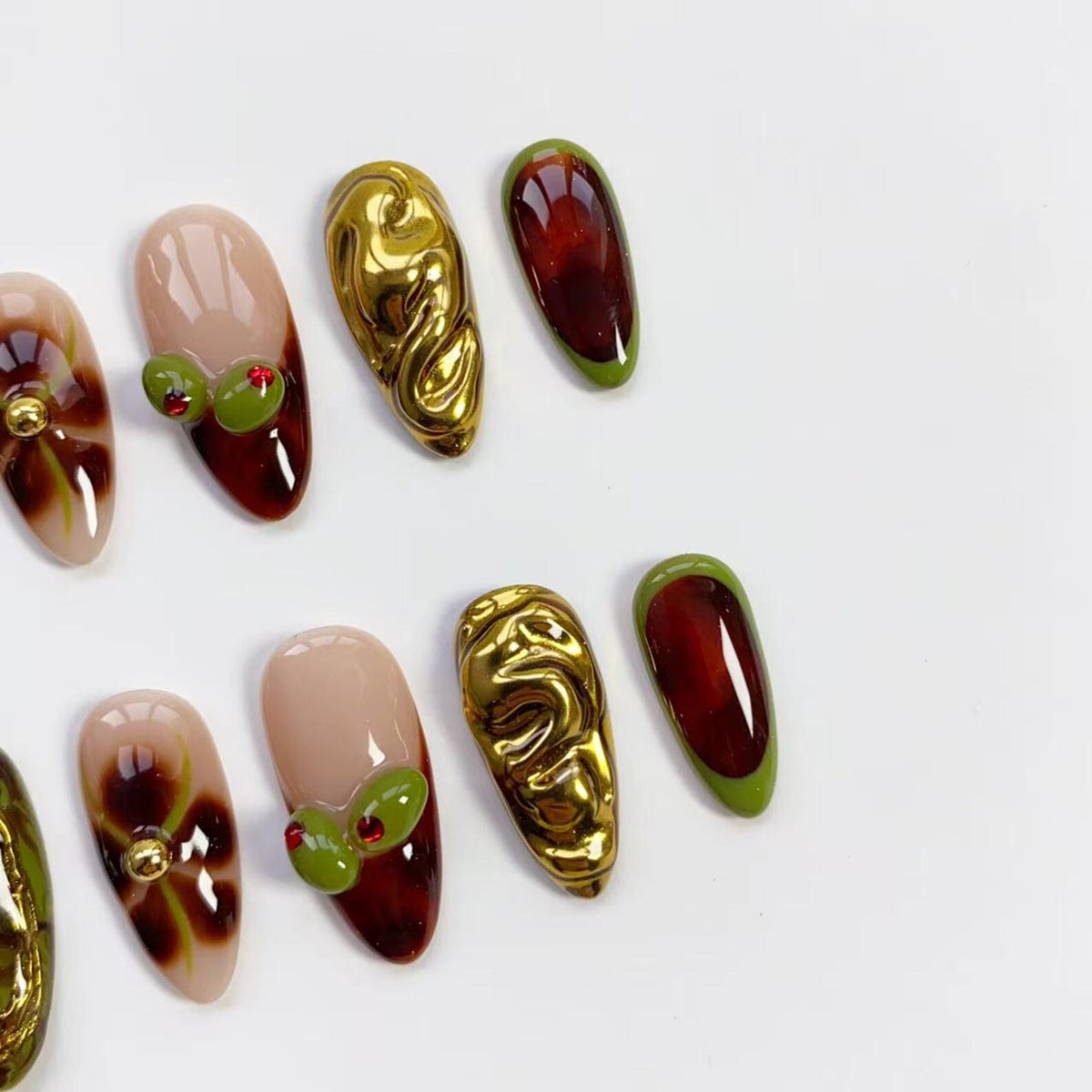 A collection of almond-shaped artificial nails in various designs. Some nails are gold, others feature a nude base with olive and red accents. Other nails are a deep red with a green border. The nails are arranged on a white surface.