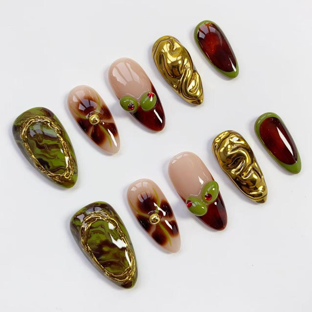 A collection of artificial nails in various designs. Some nails feature a gold swirl design, others have a marble green and gold pattern, and some have a nude base with a dark red tip and olive accents. The nails are oval-shaped.