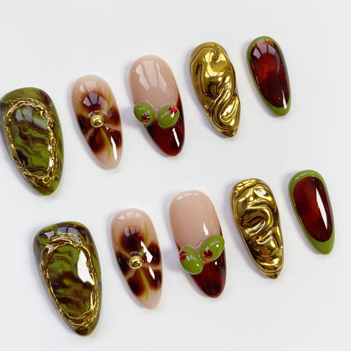 A collection of ten almond-shaped artificial nails. The nails feature various designs, including olive-like embellishments, gold metallic textures, and abstract patterns in shades of green, brown, and red. Some nails have a nude base.