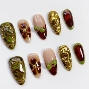 A collection of ten almond-shaped artificial nails. The nails feature various designs, including olive-like embellishments, gold metallic textures, and abstract patterns in shades of green, brown, and red. Some nails have a nude base.