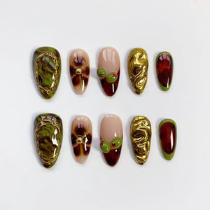 A set of ten artificial nails in various designs. Some nails feature a gold swirl design, others have olive and red accents, and some have a gold border with a green and brown marbled effect. The nails are oval-shaped.