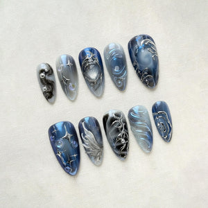 A set of ten almond-shaped artificial nails. The nails feature a variety of designs, including swirling patterns, silver embellishments, and a mix of blue, gray, and black colors. Some nails have a glossy finish.