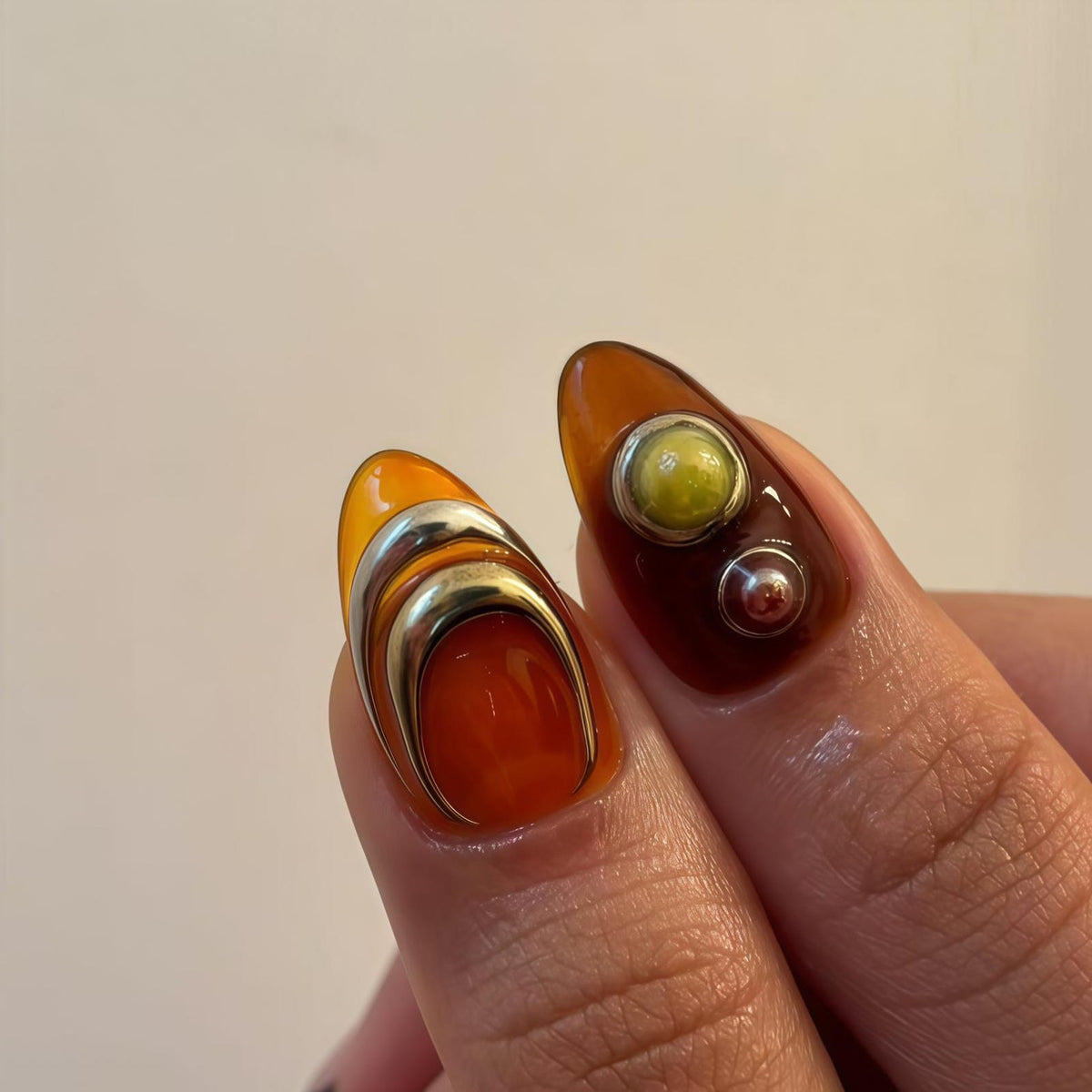 Close-up of two manicured nails. One nail features a layered silver design over orange and red polish. The other nail has a yellow-green and silver stud design over brown polish. The nails are almond-shaped.
