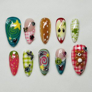 A set of ten artificial nails with various designs. The nails feature a variety of colors, including green, pink, and brown, with patterns like stars, polka dots, and plaid. Some nails have 3D embellishments such as a teddy bear and butterflies.