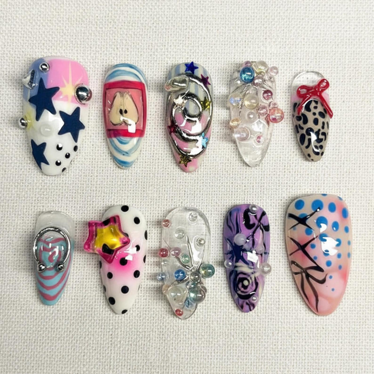 A collection of ten artificial nails with various designs. The nails feature a range of colors, including pink, blue, white, and purple, and are adorned with stars, dots, and abstract patterns. Some nails have 3D embellishments like beads and metal a