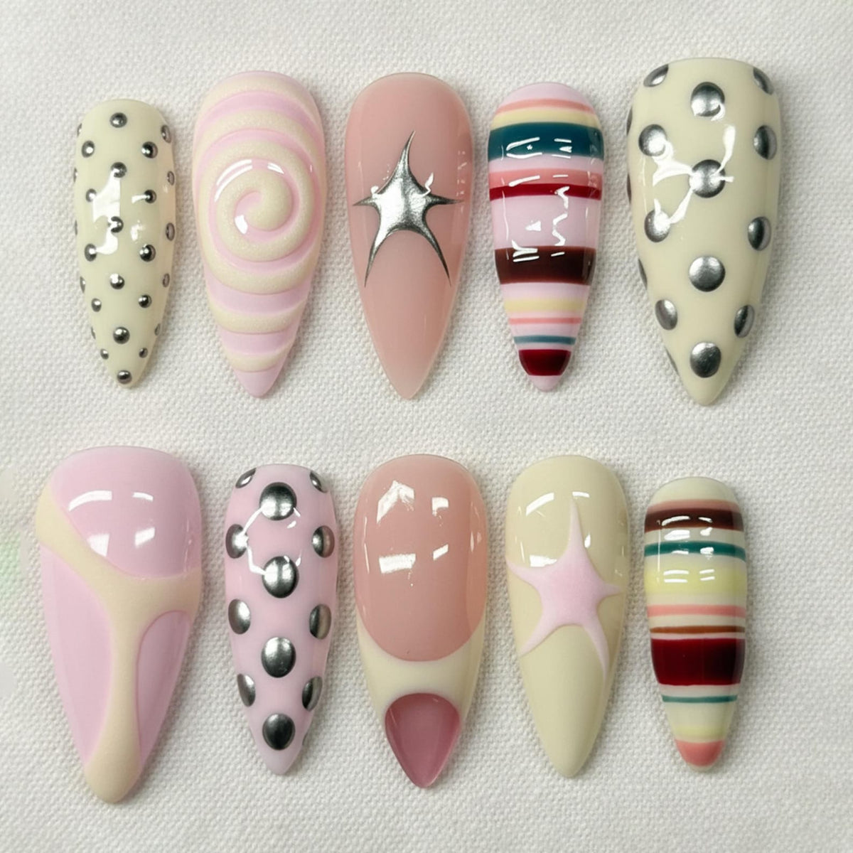 A collection of ten artificial nails in various designs. The nails feature a range of colors, including pink, cream, and silver. Some nails have a swirl pattern, while others have stripes, star accents, or silver studs. The nails are almond-shaped.