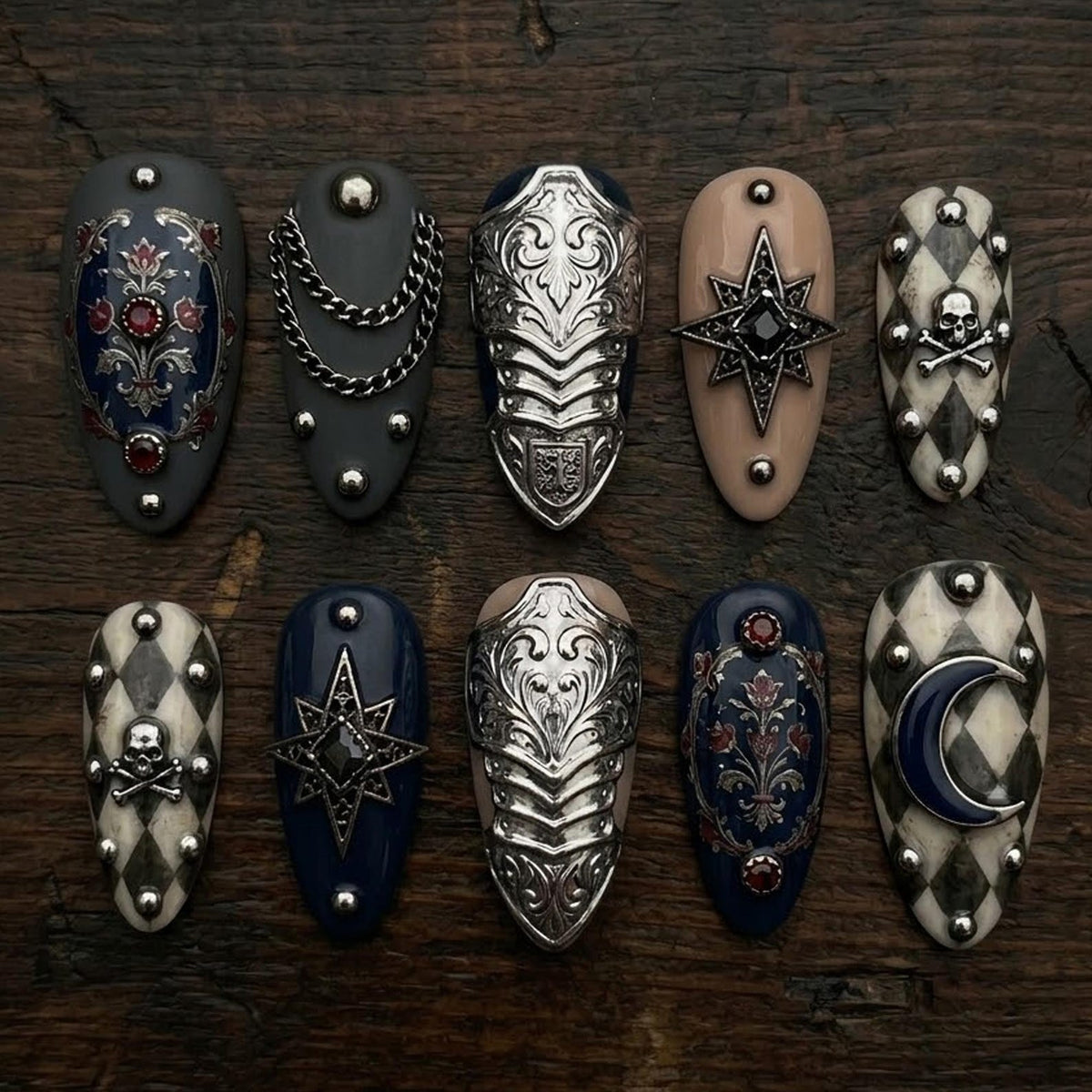 A collection of ten intricately designed nail art pieces. The nails feature various designs, including ornate silver armor, chains, starbursts, and skull and crossbones motifs. Colors include gray, navy, beige, and white, with accents of red and silv