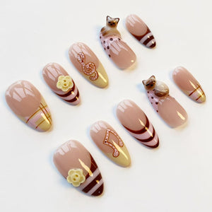 A collection of almond-shaped false nails in various designs. Some nails feature musical notes, floral accents, or cat figurines. The color palette includes nude, brown, and yellow hues, with decorative stripes and patterns.