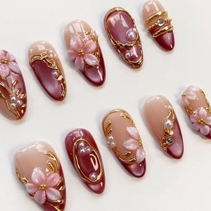 A collection of artificial nails in various designs. Some nails are a light pink with gold accents and small pearls. Others are a deep red with gold trim and pearls. Several nails feature 3D pink flower designs.