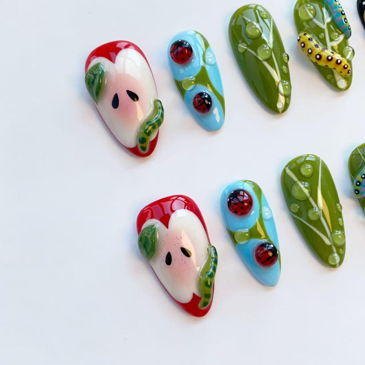A collection of artistic nail designs featuring various nature-inspired themes. Designs include a red apple with a green caterpillar, ladybugs on blue backgrounds, and green leaves with water droplets. The nails are oval-shaped and set against a whit