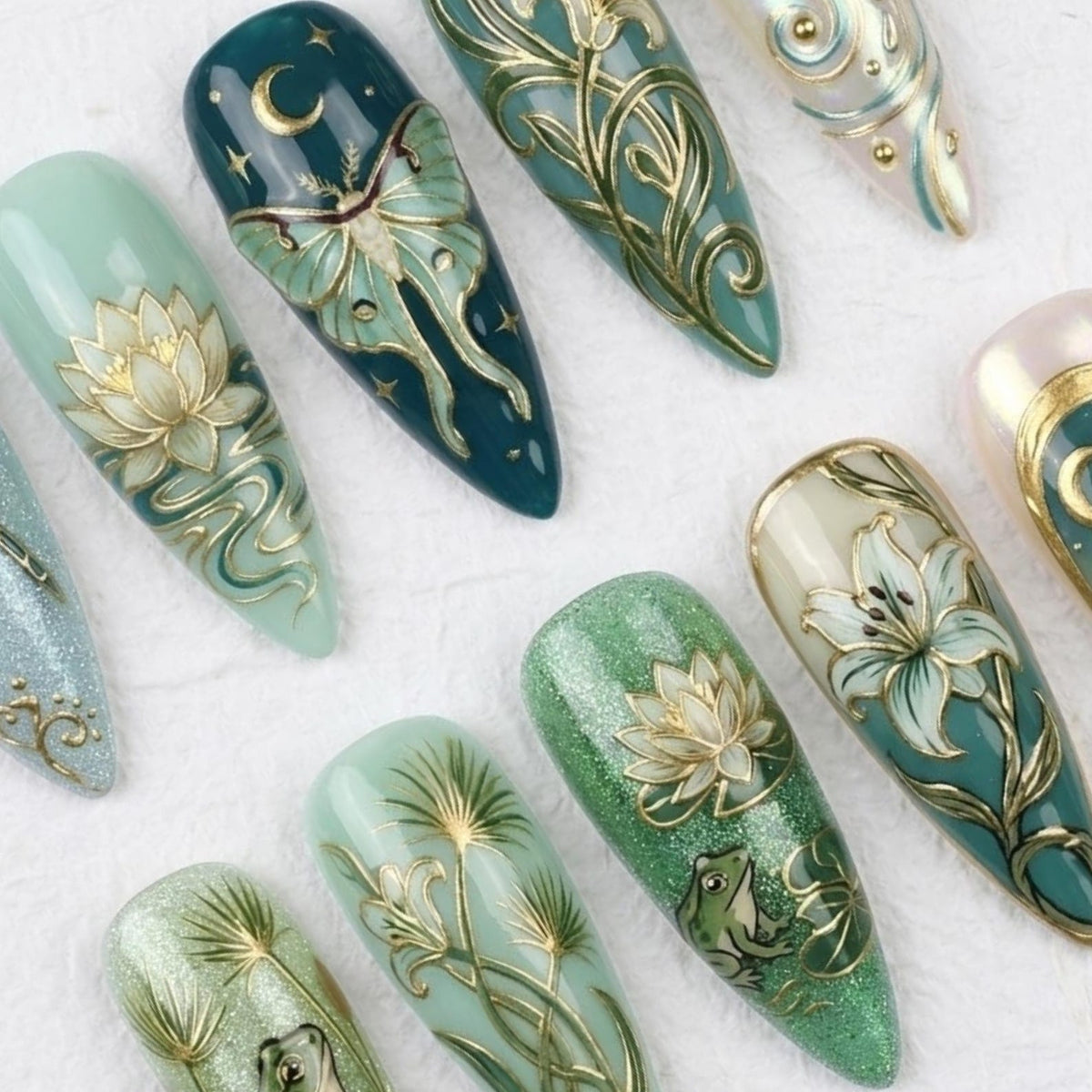A collection of long, almond-shaped artificial nails with intricate designs. The nails feature a variety of colors, including teal, green, and white, with gold accents. Designs include a moth, lotus flowers, and other botanical elements.