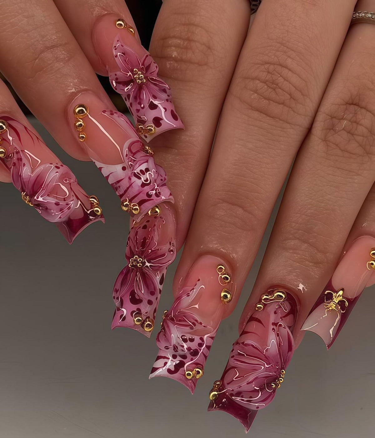 Long, sculpted fingernails with a clear base and intricate designs. The nails feature a gradient of pink and burgundy, adorned with 3D floral accents, gold beads, and heart patterns. The overall effect is elegant and artistic.