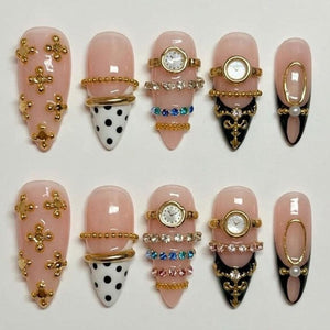 A set of ten artificial nails in various designs. Some nails are light pink with gold embellishments, while others feature watch faces, rhinestones, and black and white polka dots. The nails are almond-shaped.