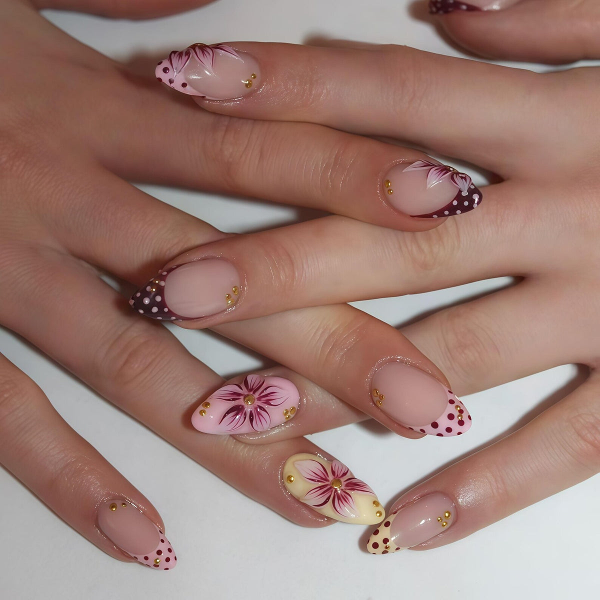 Close-up of a hand with almond-shaped nails. The nails feature a nude base with various designs, including floral patterns, burgundy polka dots, and gold accents. The overall design is elegant and artistic.
