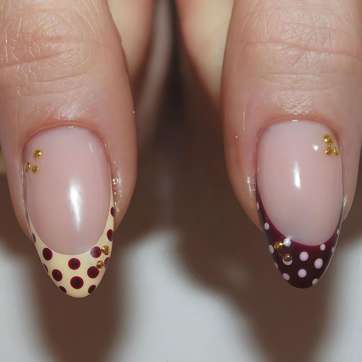 Close-up of two manicured fingernails. One nail features a cream-colored base with maroon polka dots and gold embellishments. The other nail has a maroon tip with white polka dots and gold embellishments. Both nails have a nude-colored base.