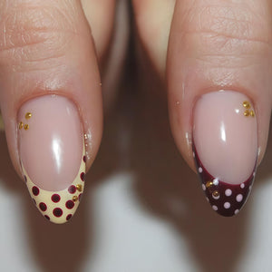 Close-up of two manicured fingernails. One nail features a cream-colored base with maroon polka dots and gold embellishments. The other nail has a maroon tip with white polka dots and gold embellishments. Both nails have a nude-colored base.