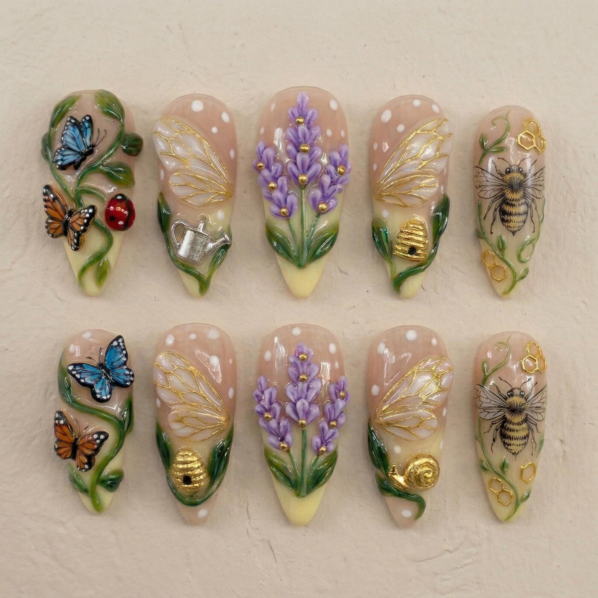 A set of ten almond-shaped artificial nails with detailed designs. The nails feature butterflies, bees, lavender, and other garden-themed elements. The base color is a light pink with white polka dots, and the designs are in various colors.
