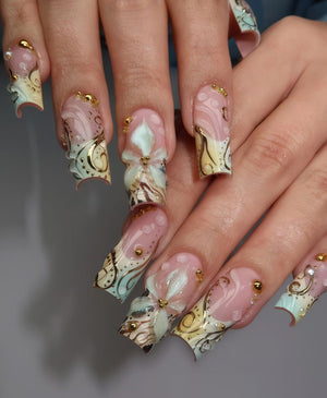 A set of long, sculpted fingernails with a light pink base and intricate designs. The nails feature gold accents, floral embellishments, and a mix of light blue and cream-colored patterns. Small gold beads are also applied.