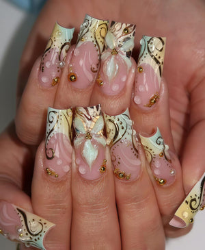 A set of long, sculpted fingernails. Each nail features a pink base with white and gold swirls, and a light blue and yellow tip. Some nails have 3D floral embellishments and gold bead accents.