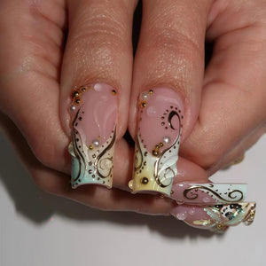 Close-up of long, decorated fingernails. The nails are painted with a light pink base and feature gold and black swirls, small gold beads, and white floral accents. The design includes a water droplet effect.