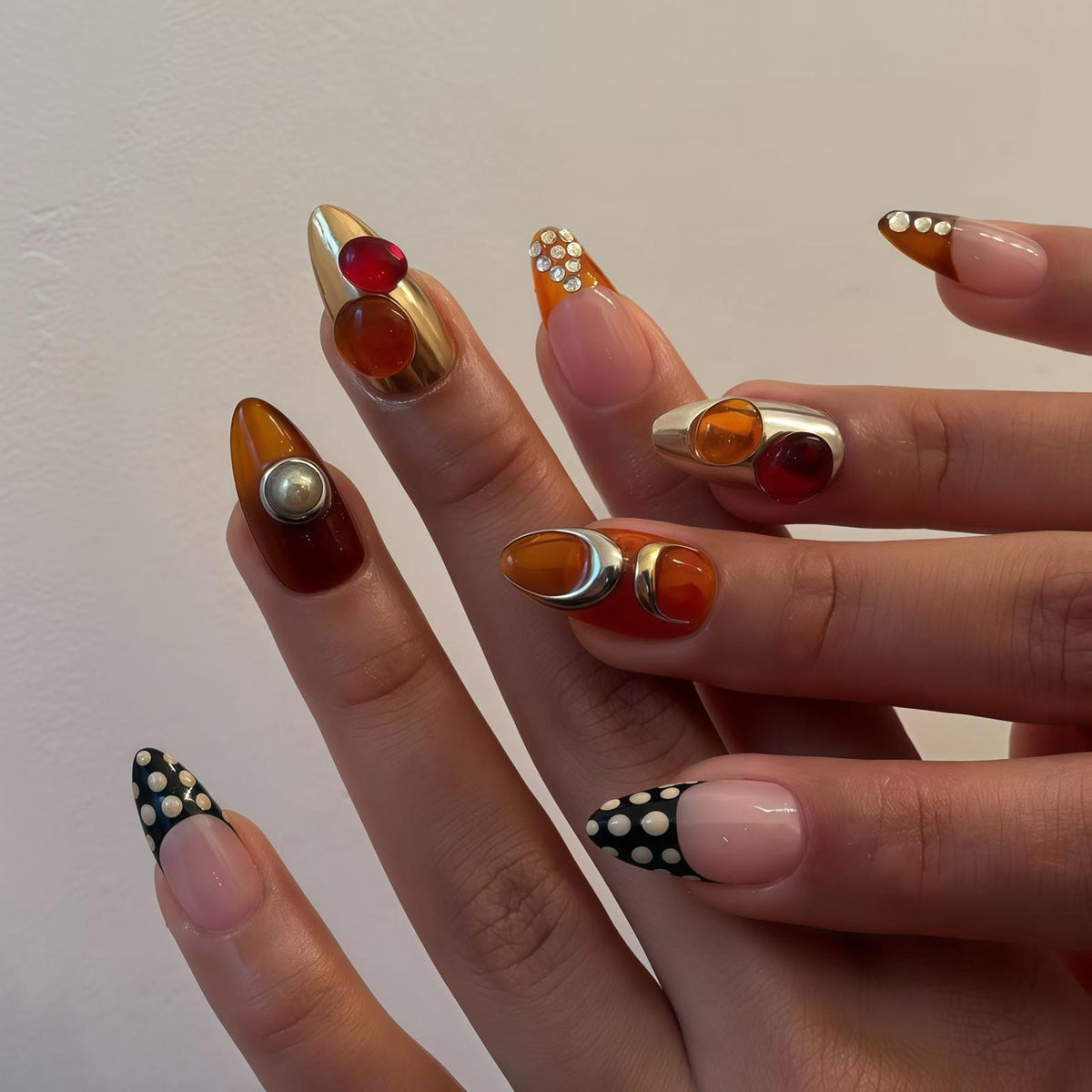 A close-up of a hand with long, almond-shaped nails. The nails feature a variety of designs, including gold metallic polish with red and orange gems, clear polish with white polka dots, and a silver ring with orange and red gems.