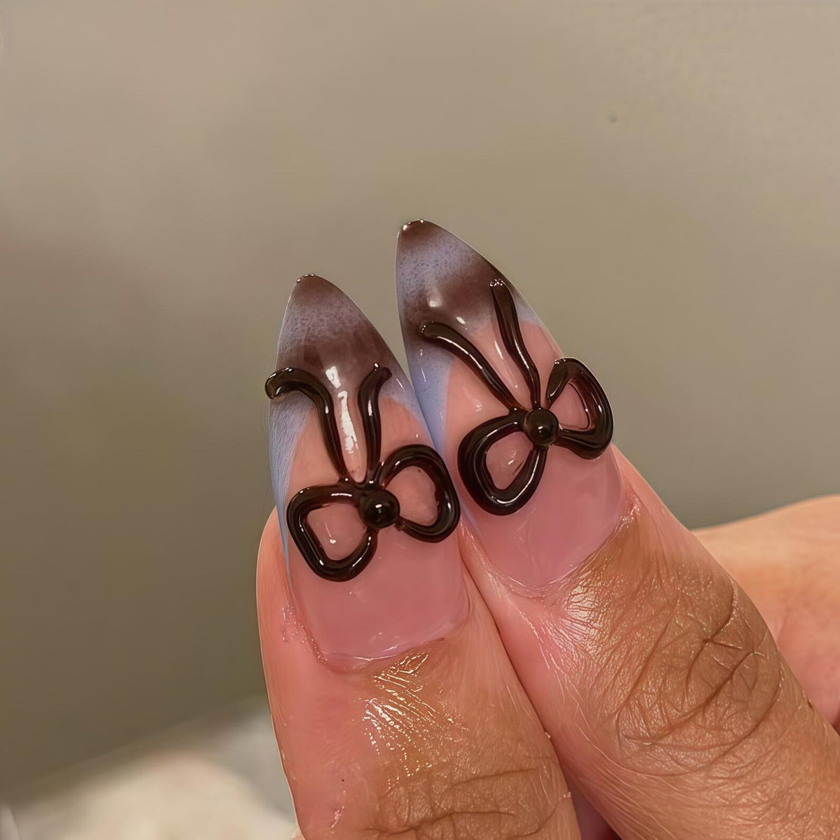 Close-up of two fingernails with a light pink base, a gradient of light blue and brown, and dark brown bow-shaped nail art. The nails are long and pointed.
