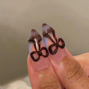 Close-up of two fingernails with a light pink base, a gradient of light blue and brown, and dark brown bow-shaped nail art. The nails are long and pointed.