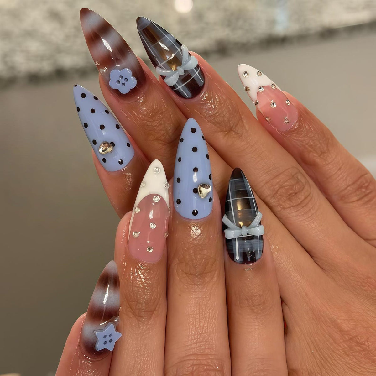 A set of long, almond-shaped artificial nails. Designs include baby blue with black polka dots, brown and white swirls, and plaid patterns. Embellishments include small bows, rhinestones, and flower and heart-shaped charms.